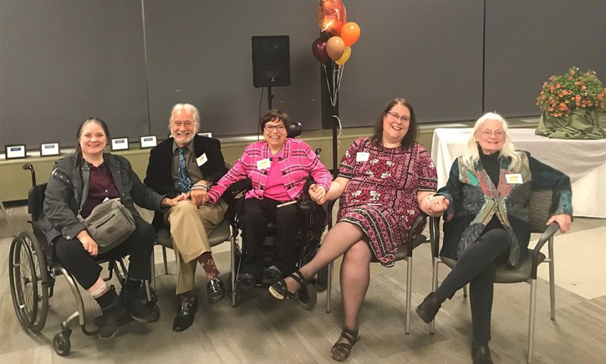 A group of past Executive Directors sits in a semi-circle holding hands.