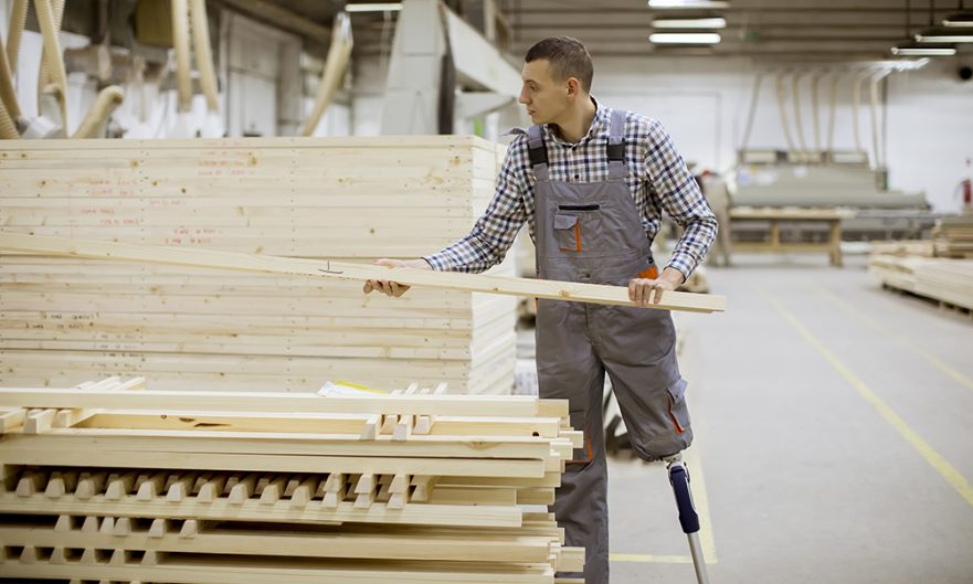 A man with a prosthetic leg lifts a large board from a pile in a lumber mill.