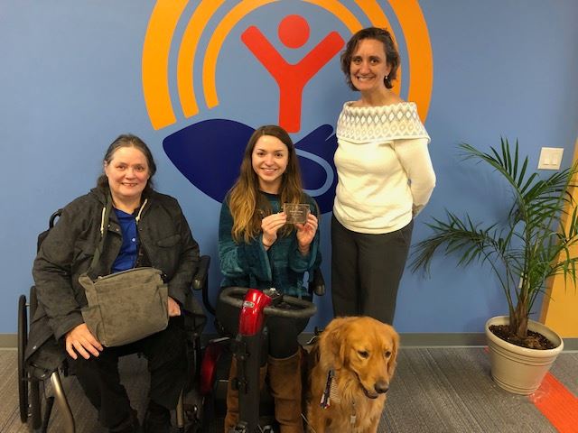 Two women sit and one stands before the camera with a guide dog.