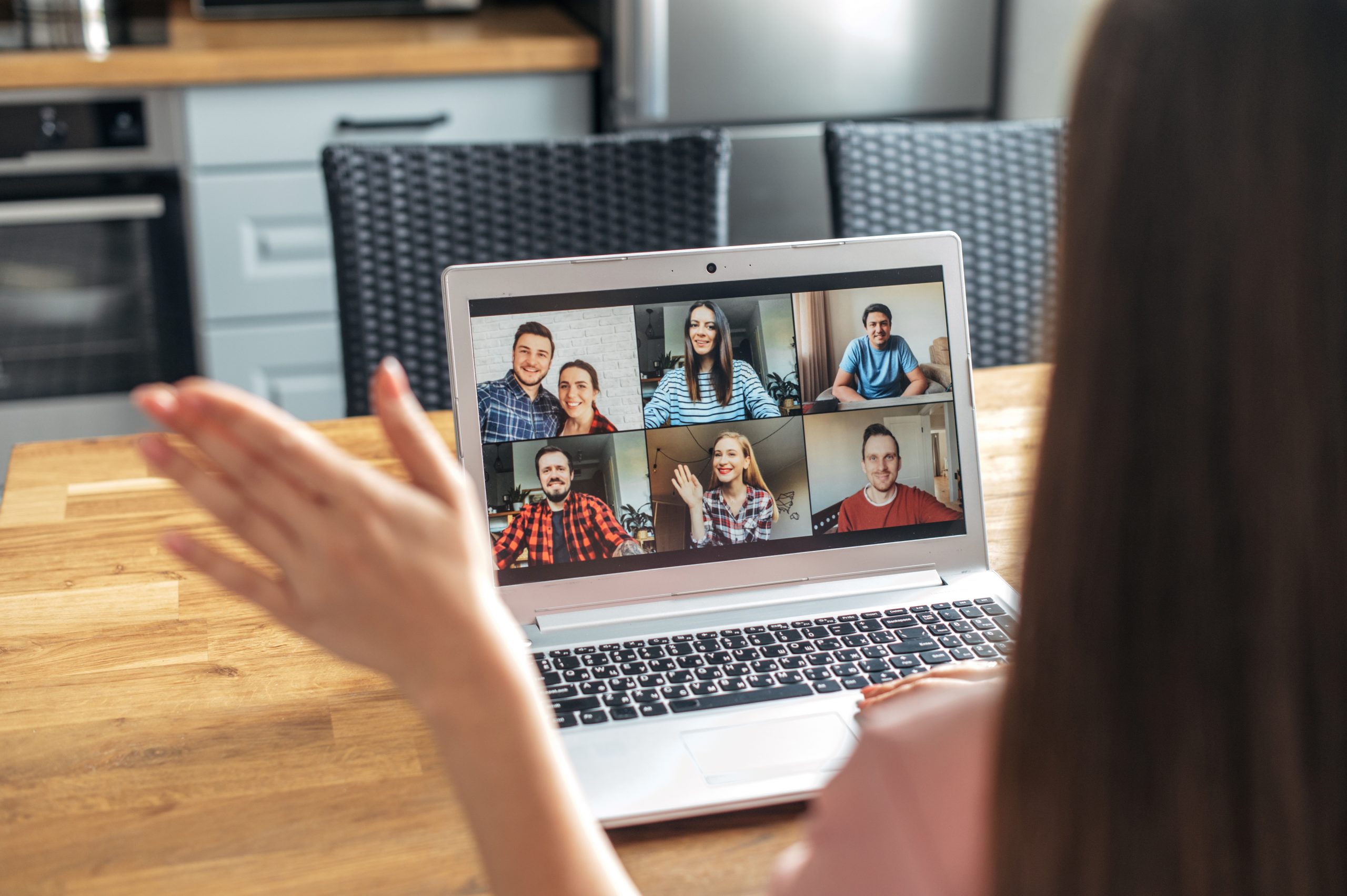 Looking over the shoulder of a young woman at her laptop screen, siting in her kitchen. The screen has 6 other people on it doing a Zoom call.