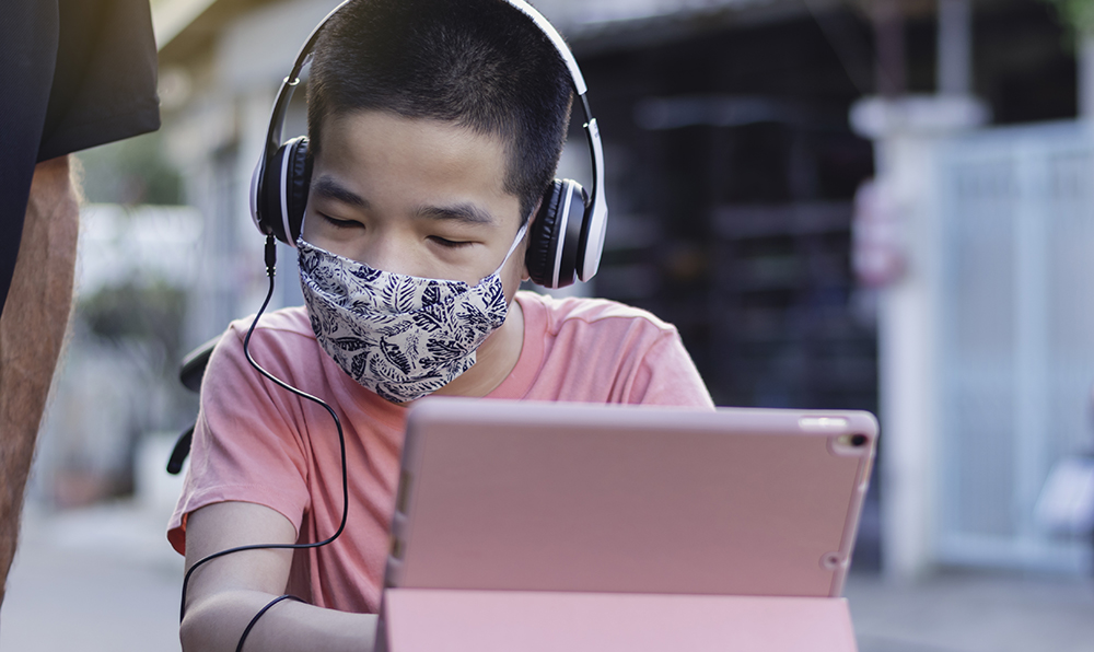 A little boy wears a COVID mask and sits in front of a tablet.