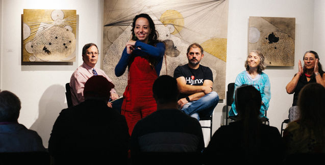 A young woman in a black shirt and red dress stands in front of a crowd using sign language.