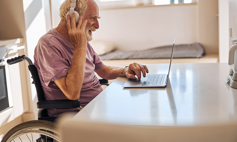 An elderly man in a wheelchair sits in an apartment in front of a laptop wearing headphones and smiling
