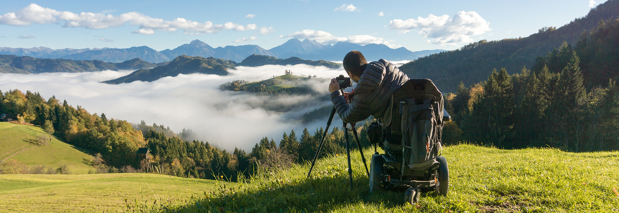 A man in a power wheelchair sits at the top of a mountain overlooking forest and other mountains covered in mist. He is using a professional camera to photograph the scene.
