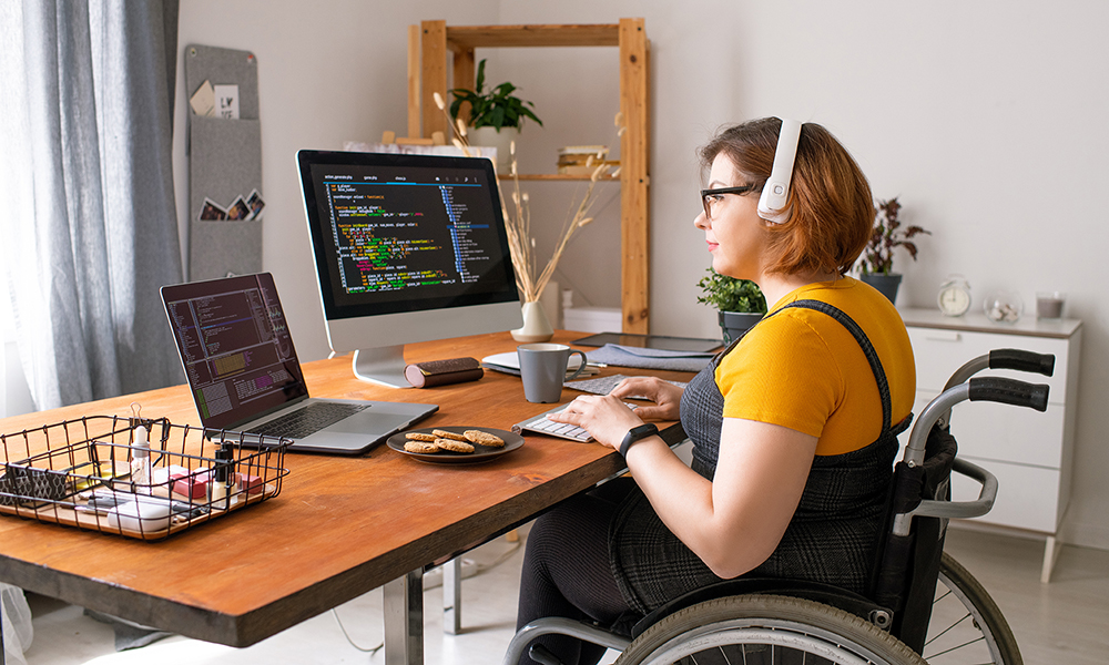 A young woman sits at a desk in her home. She is in a wheelchair in front of a computer with an additional screen, she is coding something.