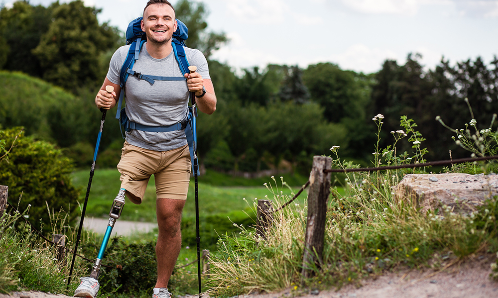 A young man with a prosthetic leg is on a hiking trail. He has a big backpack, two hiking sticks and a very happy smile.