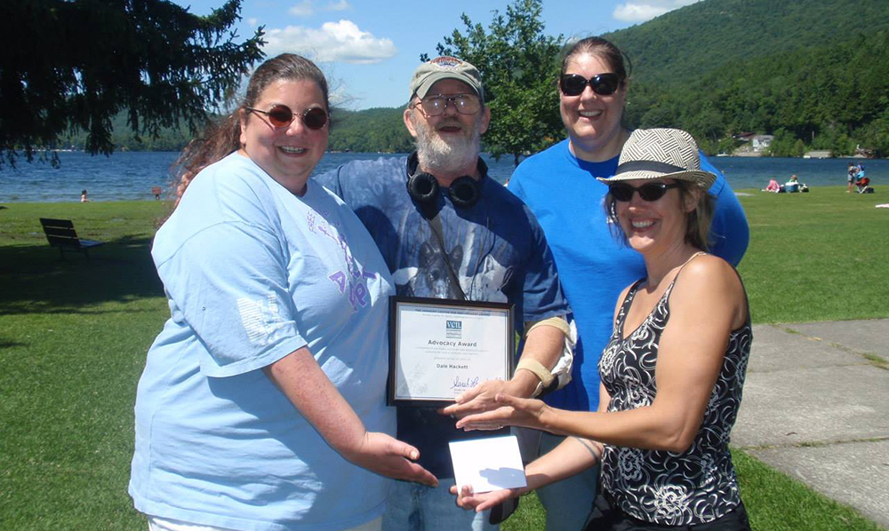 A group of VCIL staff stands in front of a pond on a sunny day, they're holding an award and all smiling.
