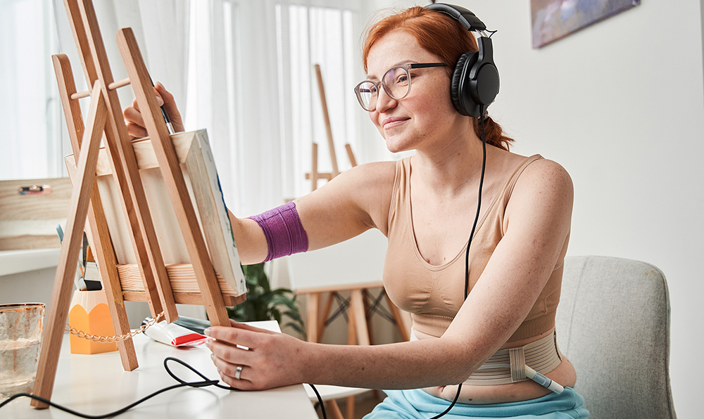 A redheaded teenage girl is painting with headphones on. She's wearing a sports bra and pants and we can see her feeding tube in her stomach.