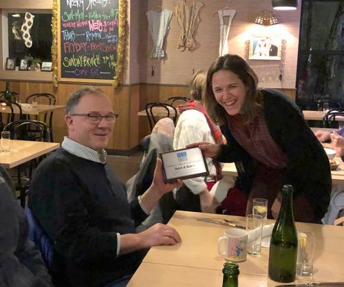 A man sits at a table as a women stands next to him presenting him with a plaque.