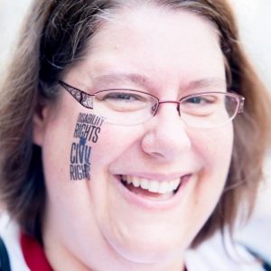 A very close up photo of Sarah Launderville. She has a big bright smile and writing on her face that says 'Disability Rights are Civil Rights.'