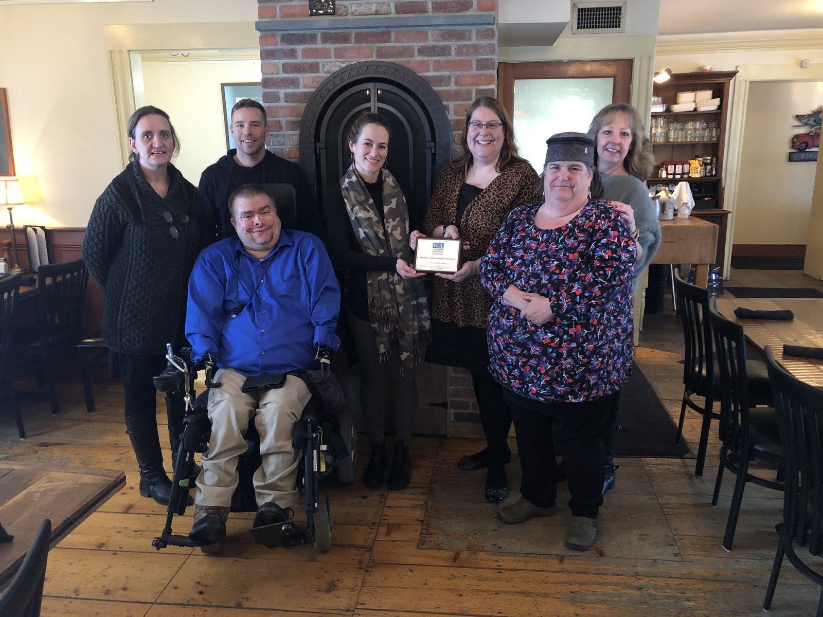 A group of people from VCIL are gathered in a group in front of a large fireplace at a restaurant, one is holding an award