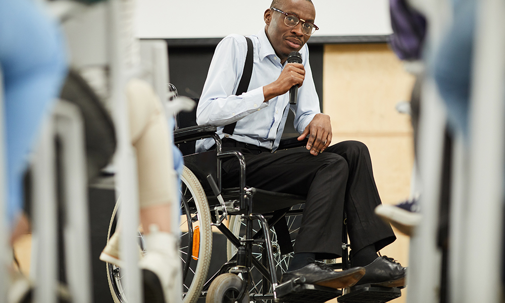 A middle aged man with glasses and cropped hair is in focus, surrounded by a classroom of people
