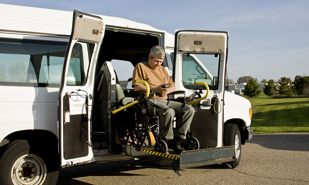A man in a chair is using the lift to get out of a van. He's in an office park on a sunny day.