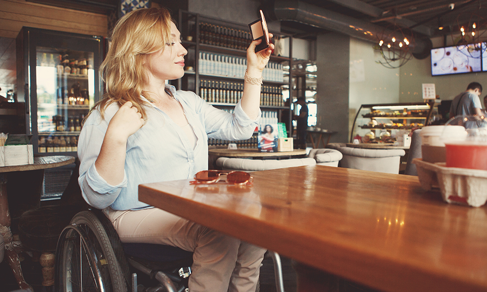 A pretty teenage girl sits in her wheelchair at a cafe. She is taking a selfie with her phone.