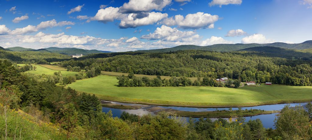 A sweeping Vermont vista on a sunny day with fluffy blue clouds in the sky and a blue stream running through.