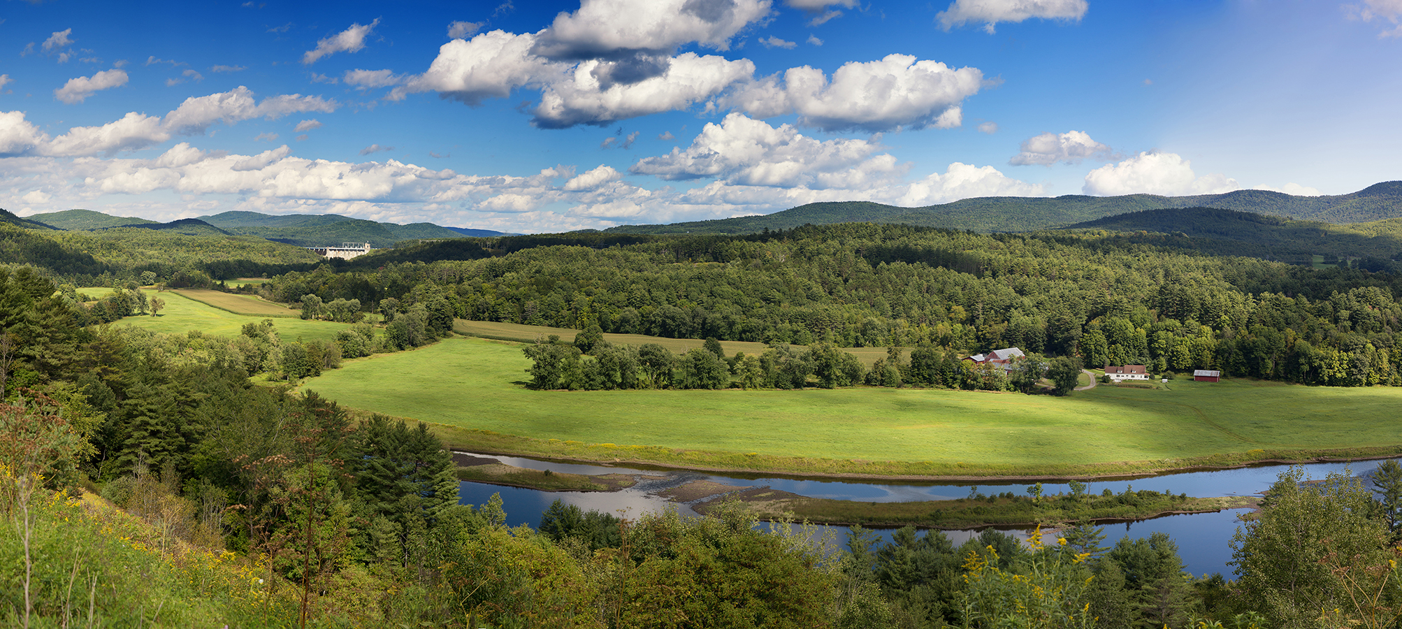 A sweeping Vermont vista on a sunny day with fluffy blue clouds in the sky and a blue stream running through.