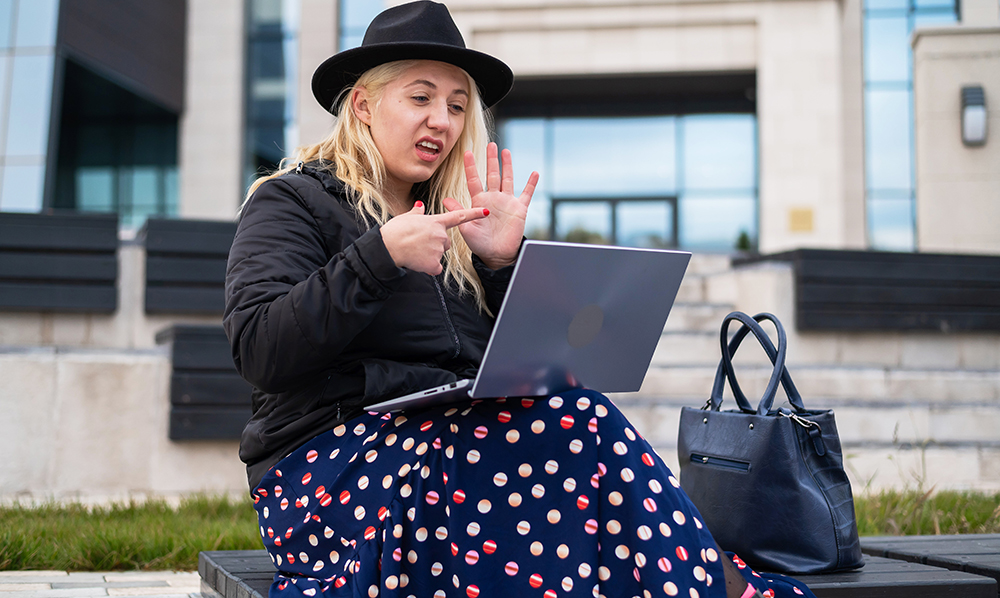 A stylish young woman is sitting in front of an office building on a bench. She is using ASL, talking to the screen of her laptop.