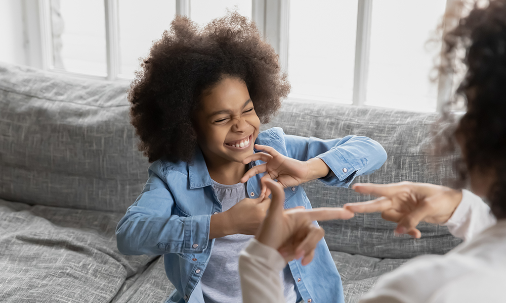 The camera looks over an adults shoulder who is using ASL with a small girl. She has a huge smile and a head full of curls