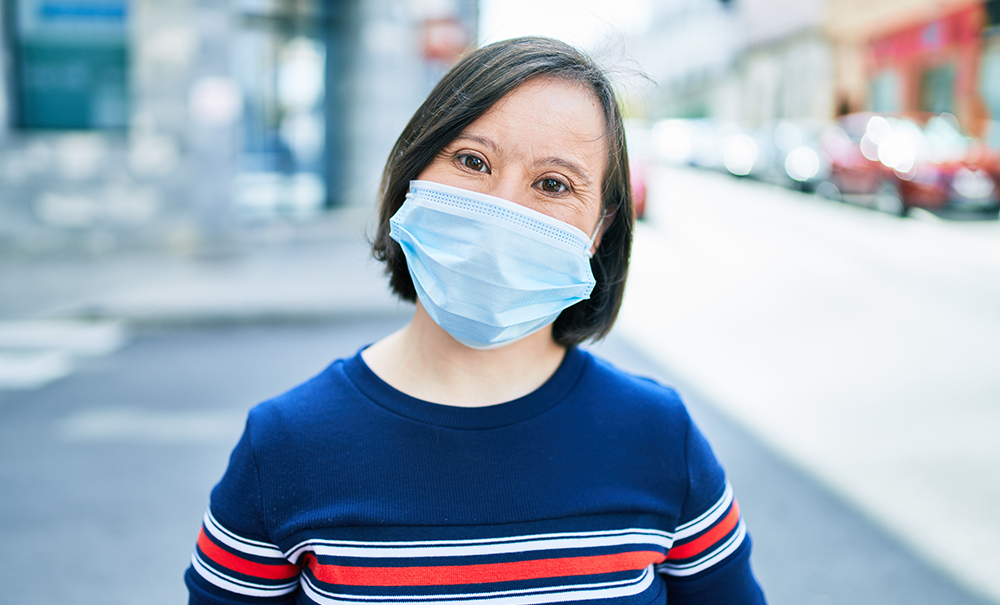 A young woman with Downs Syndrome stands directly in front of the camera wearing a protective medical mask with smiling eyes.
