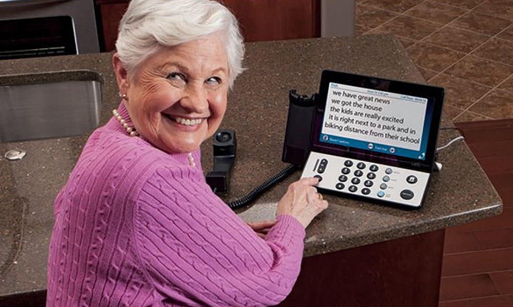 A smiling elderly woman in a pink sweater turns towards the camera from her caption phone