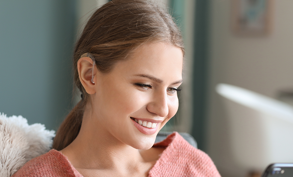 A smiling young woman with a hearing aid is sitting casually looking at her mobile phone