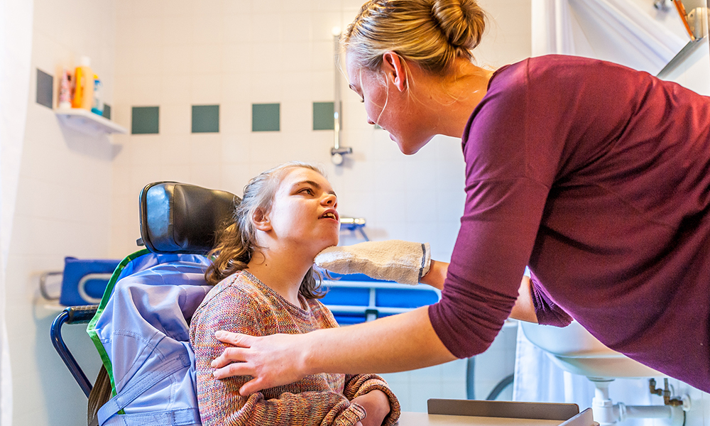 A blonde woman is leaning down and assisting a young girl in a wheelchair. She is cleaning her in a bathroom.