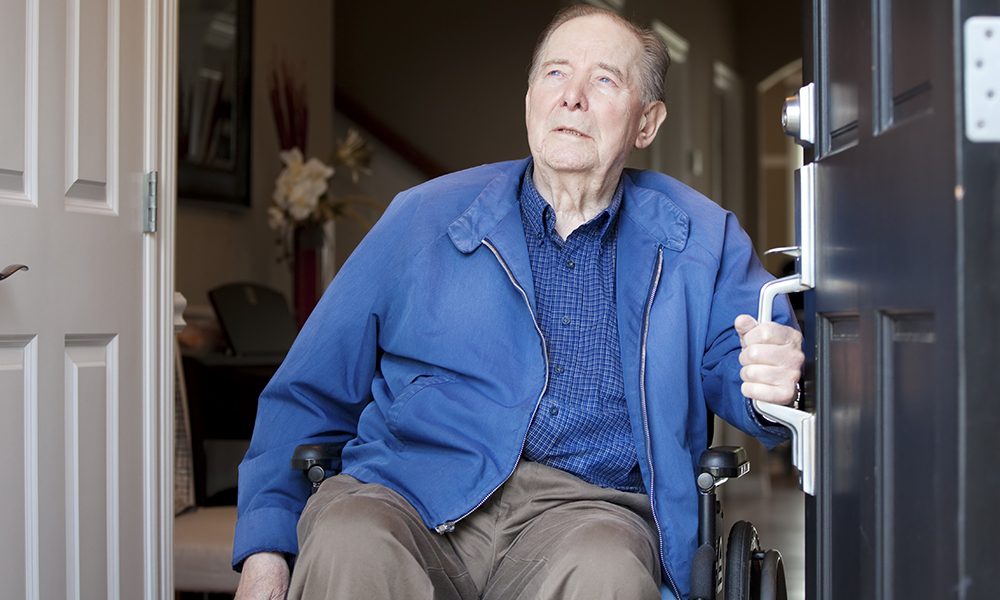 An older man in a power chair wearing a blue jacket is exiting his front door.