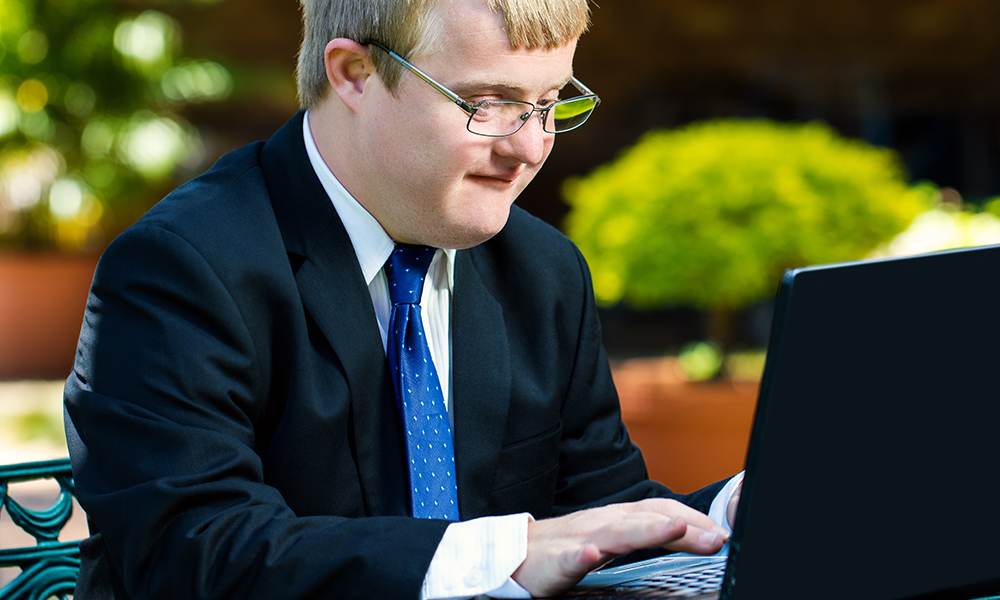 A young adult man is wearing a suit and blue tie. He has Downs Sydrome and is sitting at a laptop very focused.