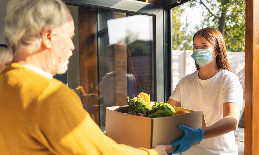 A young girl is delivering a box of groceries to an older man at his front door.