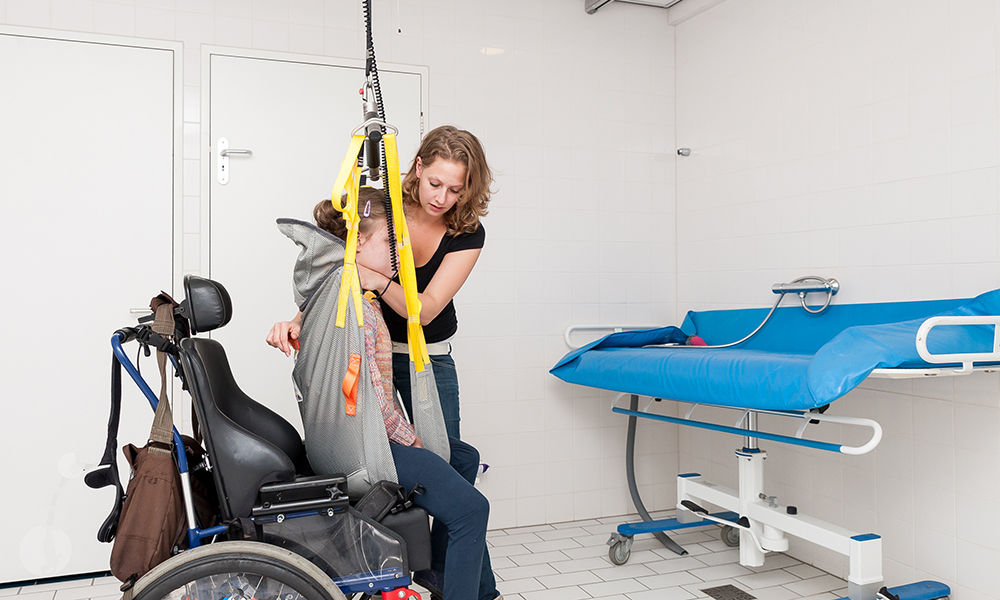 A young girl in a power chair is being assisted to a table for bathing by a blonde woman