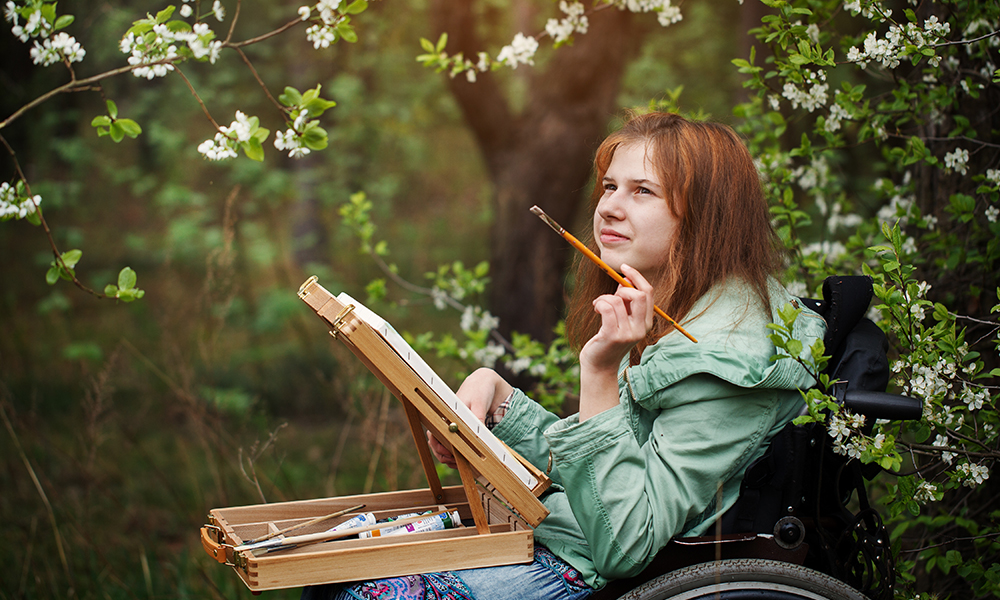 A young redheaded woman in a power chair sits amongst flowering trees with a painting palette and brush