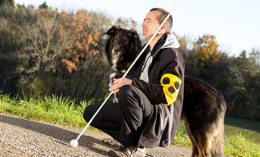 A man who is blind is kneeling down besides his guide dog. The dog is large and black and looks towards the camera