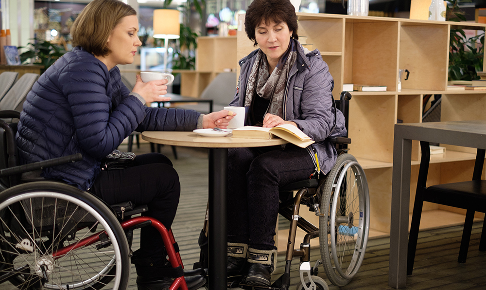 Two women in wheelchairs sit around a small table in a bookstore, both looking at the same book