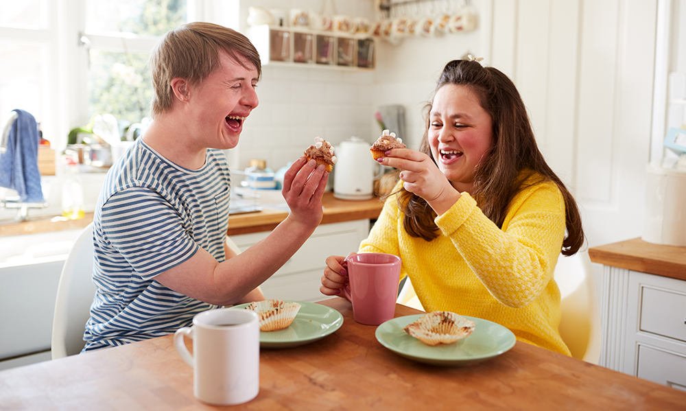 A young adult couple with Downs Syndrome are laughing in a bright cheery kitchen, they are have a 'cheers' with cupcakes