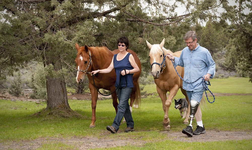 A middle aged man with a prosthetic leg and a woman walk two horses through a tree lined path.