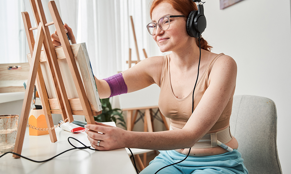 A redheaded teenage girl is painting with headphones on. She's wearing a sports bra and pants and we can see her feeding tube in her stomach