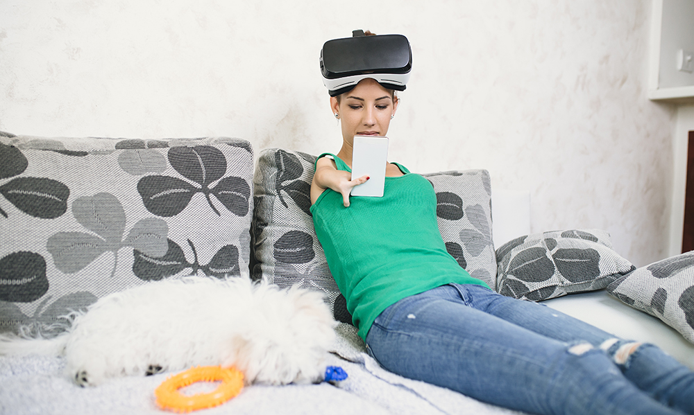 A teenage girl sits in bed with her small white dog. She's wearing a VR headset and has a limb difference on her arm