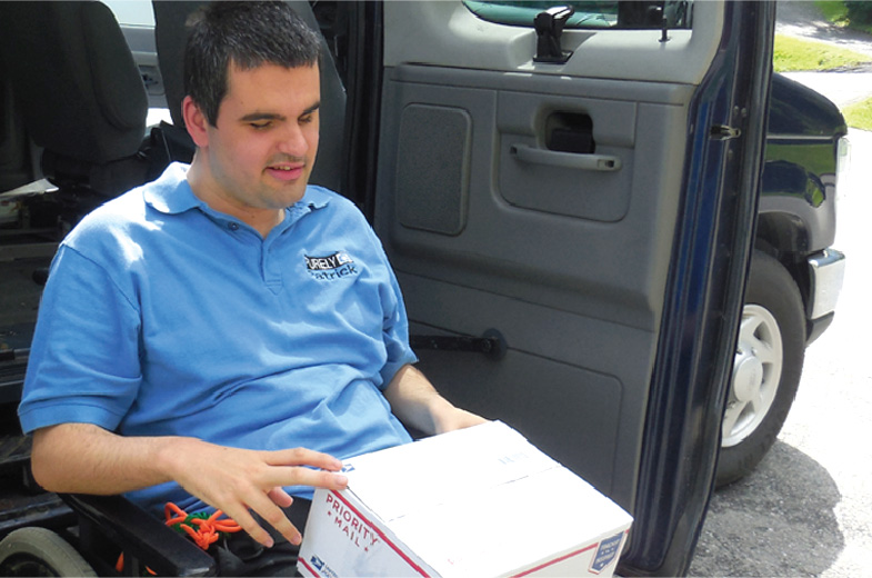 A young man in a wheelchair getting out of a van with a postal service box in his lap.